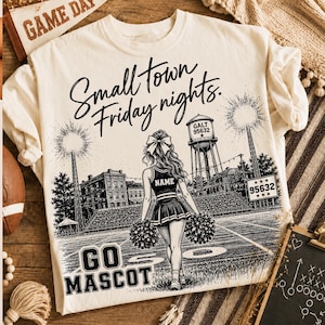 May include: A cream-colored t-shirt featuring a black graphic of a cheerleader on a football field with the text "Small town Friday nights." and "GO MASCOT". The design includes a water tower labeled "GALT 95632" and a sign reading "95632".