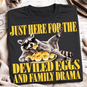 May include: A black t-shirt featuring a graphic of a raccoon wearing sunglasses, eating a deviled egg, and holding a plate of deviled eggs. The text reads: JUST HERE FOR THE DEVILED EGGS AND FAMILY DRAMA in bold yellow capital letters.