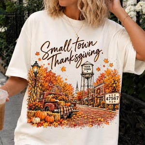 Op de afbeelding: Een off-white t-shirt met een grafische print van een herfstachtig stadje en de tekst "Small Town Thanksgiving." Het ontwerp bevat een rode truck, pompoenen, een watertoren met "YOUR TOWN" en een bord met het nummer "47567."