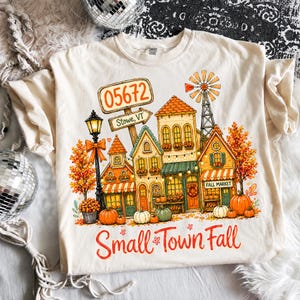 Pode incluir: Uma t-shirt de cor creme com um gráfico colorido de uma pequena cidade no outono. O design inclui edifícios, um moinho de vento, abóboras, árvores com folhas cor de laranja, um candeeiro de rua e um sinal com 05672 Stowe, VT. O texto Small Town Fall está impresso na parte inferior.