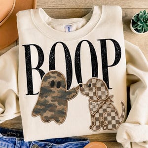 Peut inclure: Un sweat-shirt à col rond couleur crème avec le texte noir vieilli BOOP. Sous le texte, un fantôme à motif camouflage touche le nez d'un chien à carreaux marron et blanc portant un bandana assorti à carreaux.