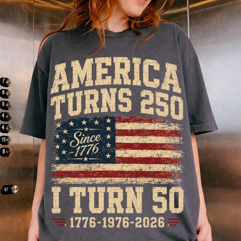May include: Dark gray t-shirt with the text "AMERICA TURNS 250" in a distressed, vintage style. Below is a distressed American flag graphic and the text "I TURN 50". The shirt also includes the text "Since 1776" and "1776-1976-2026".