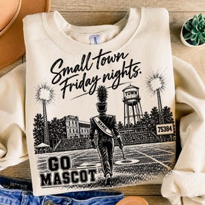 May include: A cream-colored crewneck sweatshirt featuring a black graphic print of a marching band member on a football field. The text reads Small town Friday nights, TOWN, 75384, GO MASCOT, and NAME on the band member's sash.