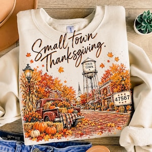Könnte beinhalten: Ein cremefarbenes Sweatshirt mit Rundhalsausschnitt und einer Grafik einer herbstlichen Kleinstadt mit rotem Pickup, Kürbissen, einem Wasserturm mit der Aufschrift "YOUR TOWN" und einem Schild "47567". Darüber steht "Small town Thanksgiving".