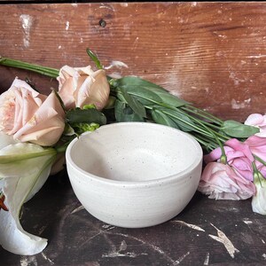 May include: A small, round, off-white ceramic bowl with a speckled texture and a smooth, curved rim. The bowl features a simple, minimalist design with a matte finish, showcasing subtle variations in the clay surface and a consistent, rounded shape.