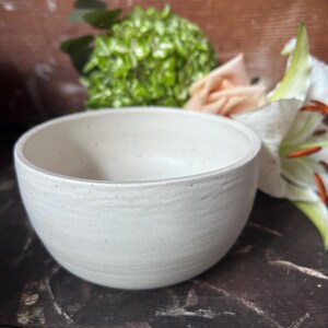 May include: A round, off-white ceramic bowl with a speckled texture and subtle horizontal ridges along the exterior surface. The bowl features a smooth, matte finish and a simple, clean design with a wide opening and a rounded base.
