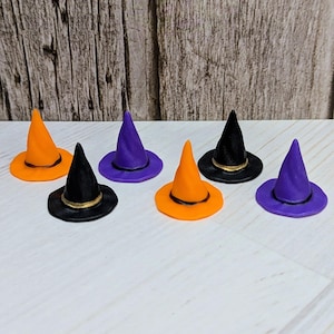 May include: A collection of six miniature witch hats in orange, purple, and black colors. Each hat features a pointed crown and a wide brim, with a thin band encircling the base of the crown. The hats are arranged in two rows on a light surface.
