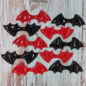 May include: A collection of glossy, bat-shaped appliques in red and black. The appliques are arranged on a light wood-grain surface. The bat wings are detailed with veining, and the overall design has a shiny, reflective quality.