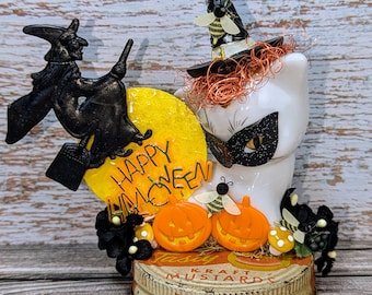 May include: A Halloween-themed decoration featuring a white cat wearing a black mask and witch hat, with a yellow moon and the words "Happy Halloween." Includes orange pumpkins, a black witch on a broom, and a bee. The base is a Kraft Mustard lid.