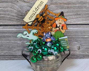 May include: A silver cauldron filled with green orbs and googly eyes, topped with Halloween-themed decorations. Includes a witch, ghost, spider web, and a sign that says "cauldron". A small book reads "Spells, Curses, Potions".