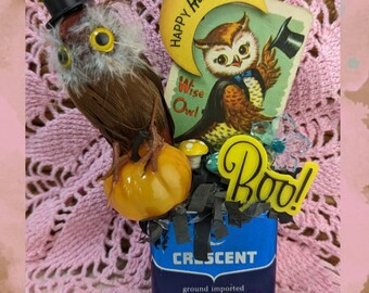 May include: A Halloween-themed arrangement featuring a brown owl wearing a top hat, perched on a small orange pumpkin. A vintage card with a cartoon owl and the words "Happy Halloween" and "Wise Owl" is included, along with a "Boo!" sign.