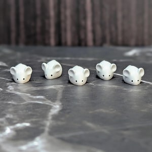 May include: Five small, white ceramic mouse figurines arranged in a horizontal row. Each mouse features a smooth, rounded body, two prominent ears, and two tiny black dots representing eyes, all crafted with a clean, matte white finish.