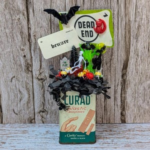 May include: A Halloween-themed display featuring a vintage Curad bandage tin. The tin is decorated with miniature Halloween figures, including bats, spiders, and a graveyard scene. The words "DEAD END" and "beware" are displayed on signs.