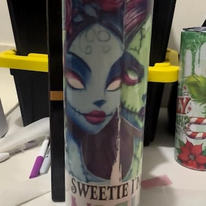Puede incluir: Un vaso cil&iacute;ndrico con una ilustraci&oacute;n espeluznante de un personaje con piel azul, labios rojos y boca cosida. El vaso tiene el texto "Sweetie Night" impreso. El dise&ntilde;o es una mezcla de azul, verde y morado.