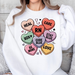 May include: White sweatshirt featuring a colorful graphic of candy hearts with medical-themed phrases like "HEAL," "CARE," and "RN." The design includes a stethoscope, syringe, and a heartbeat line. A cute and cozy top for nurses.