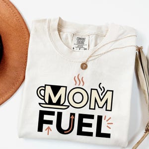 Mom Fuel retro coffee typography graphic t-shirt printed on Comfort Colors garment-dyed vintage washed heavyweight tee featuring coffee cup M letterform with steam lines and energy arrows in terracotta black and cream