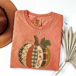 Könnte beinhalten: Ein kurzärmeliges T-Shirt in Terrakotta mit einer großen Kürbisgrafik, die aus verschiedenen stoffartigen Flicken wie Karomustern, Blumenmustern und einfarbigen Stoffen besteht. Das T-Shirt hat einen Rundhalsausschnitt mit einem Comfort Colors Etikett.