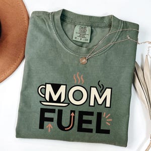 Mom Fuel retro coffee typography graphic t-shirt printed on Comfort Colors garment-dyed vintage washed heavyweight tee featuring coffee cup M letterform with steam lines and energy arrows in terracotta black and cream