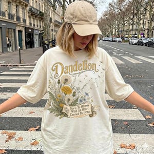 May include: A cream-colored short-sleeve t-shirt featuring a graphic of yellow and white dandelion flowers with the text "Dandelion" and "Born To Bloom Wild At Heart Carried By Grace" on a banner, paired with a solid beige baseball cap.