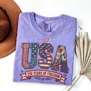 May include: A lilac-colored t-shirt with the word "USA" in a patchwork design featuring red gingham, denim, and floral patterns. The shirt also has the text "250 YEARS OF FREEDOM" on a banner.