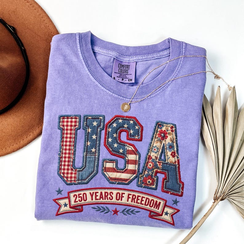 May include: A lilac-colored t-shirt with the word "USA" in a patchwork design featuring red gingham, denim, and floral patterns. The shirt also has the text "250 YEARS OF FREEDOM" on a banner.