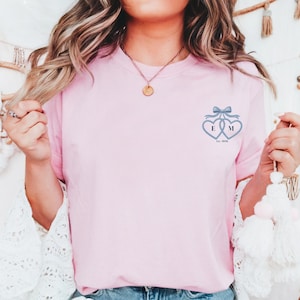 May include: A pale pink t-shirt with a blue embroidered design of two hearts linked together, with the letters "E" and "M" inside. A blue bow sits above the hearts, and the text "Est. 2020" is below.