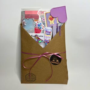 May include: A brown paper envelope tied with a pink ribbon, containing stationery items. The envelope is sealed with a wax seal and a sticker that says "Book Review". Inside are stickers, a purple heart-shaped bookmark, and other decorative elements.