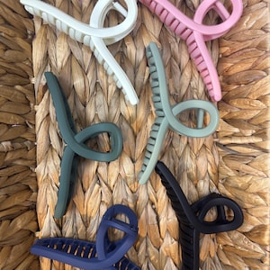 Puede incluir: Colección de seis pinzas para el cabello en forma de garra, en varios colores: blanco, rosa, verde, azul y negro. Las pinzas de plástico tienen un diseño curvo con dientes para sujetar el cabello. Dispuestas sobre una cesta tejida.