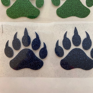 May include: Four glitter bear paw prints, two green and two blue. The paw prints are cut out and ready to be ironed onto fabric.