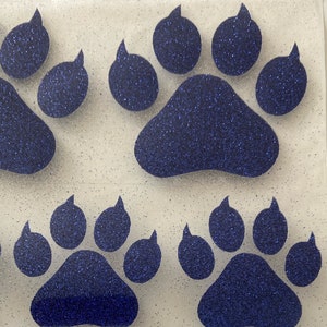 May include: Three blue glitter paw prints on a white background. The paw prints are in different sizes and positions.