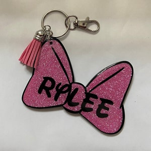 May include: Pink glitter bow keychain with a black outline and the name "Rylee" written in black. The keychain has a pink tassel attached to a silver key ring.