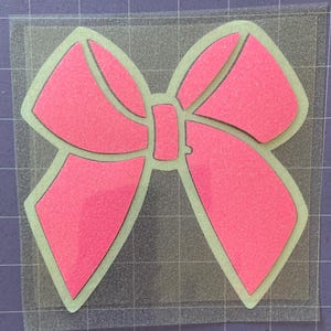 May include: A pink glitter bow with a simple design. The bow is cut out of a sheet of glitter paper and is ready to be used for crafting projects.