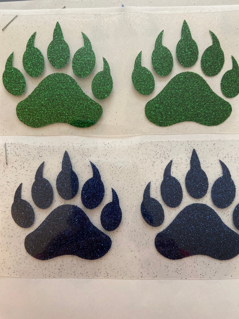 May include: Four glitter bear paw prints, two green and two blue. The paw prints are cut out and ready to be ironed onto fabric.