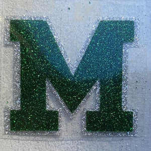 May include: A green glitter letter M with a white outline. The letter is on a clear background.