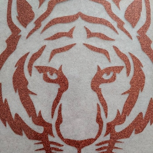May include: A close-up of a copper glitter tiger head silhouette. The tiger's face is outlined in a bold, copper glitter design, with a detailed nose, whiskers, and eyes.