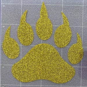 May include: A gold glitter bear paw print with five toes. The paw print is on a gray background.