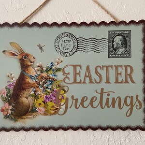 May include: A vintage-style Easter greeting sign with a brown bunny rabbit holding a basket of flowers. The sign has a blue background with a brown border and the text "Easter Greetings" in gold lettering.