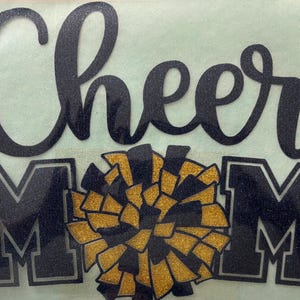 May include: Black glitter text on a light green background reads "Cheer Mom" with a gold and black pom-pom graphic. The letters "MOM" are in black with a silver outline.