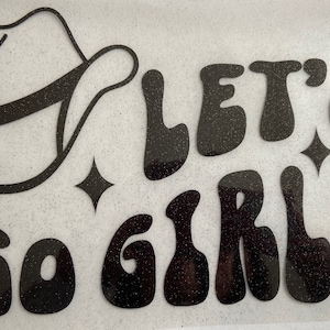May include: Black glitter iron-on design with the text "Let's Go Girls" in a retro font. A black glitter cowboy hat is in the upper left corner.