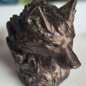 Può includere: Una statuetta a forma di lupo color marrone scuro con una finitura metallica e sottili accenti glitterati. La scultura presenta texture del pelo dettagliate, orecchie a punta e un muso rivolto in avanti, raffigurante testa e collo.