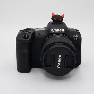 May include: Black Canon EOS R5 camera with a lens cap and a small devil figurine on top. The camera has the Canon logo and EOS R5 model name printed on it. The devil figurine is black with red horns.