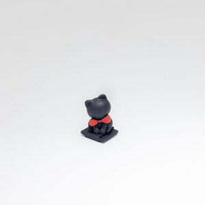 May include: A small, black cat figurine with a bright red bow tie. The cat is sitting on a small, dark square base. The figurine has a smooth, matte finish and is set against a plain white background.