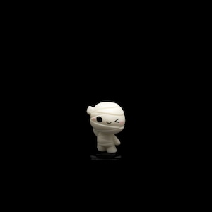 May include: A small, white figurine of a mummy with a cute, cartoonish design. The mummy has bandages around its head and body, with black eyes and a small, smiling mouth. The figurine is standing on a black surface against a black background.