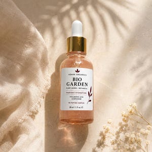 K&ouml;nnte beinhalten: Eine Glasflasche Adara Organics Bio Garden Serum mit goldener Tropferspitze. Das Etikett lautet "Plant Based Botanical Radiant Hydration" und listet Inhaltsstoffe auf. Die Flasche enth&auml;lt 30 ml Produkt. Der Hintergrund umfasst ein beiges Tuch und Trockenblumen.