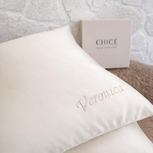 May include: Ivory-colored pillow with the name "Veronica" embroidered in a delicate script. A box with the word "CHICÉ" and other text is visible in the background. The pillow is likely part of a bedding set.