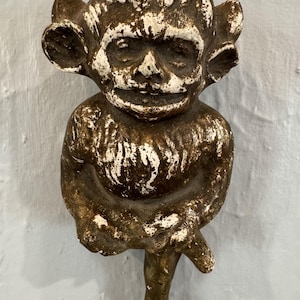 May include: A detailed, antique-style figurine of a goblin-like creature. The sculpture is primarily brown with white accents, featuring large ears, a wide grin, and a seated pose. The aged appearance suggests it is a vintage or weathered piece.