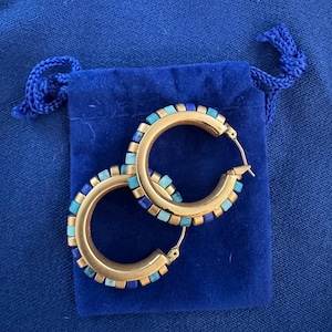 May include: A pair of gold-tone hoop earrings featuring a series of small, alternating square-shaped inlays in shades of light blue, turquoise, and dark blue. The earrings have a hinged post closure and are displayed on a blue velvet drawstring pouch.