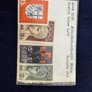 May include: A collection of vintage Saar stamps from 1956, displayed in a clear protective sleeve. The stamps feature various designs, including a coat of arms, Olympic rings, and classical figures. The text "SAAR 1956" is visible.