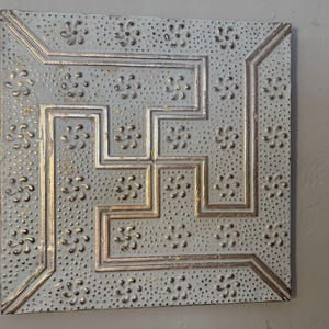 May include: Square, light gray metal wall decor with a raised, geometric design. The design features a series of stepped lines and floral accents, all in a metallic gold color. The surface has a textured, dotted pattern.
