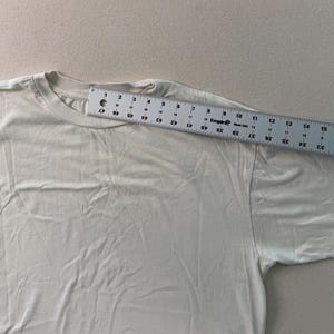 May include: A cream-colored t-shirt with a crew neck. A ruler is placed across the shoulder of the shirt, showing measurements in centimeters. The shirt is made of a soft, plain fabric.